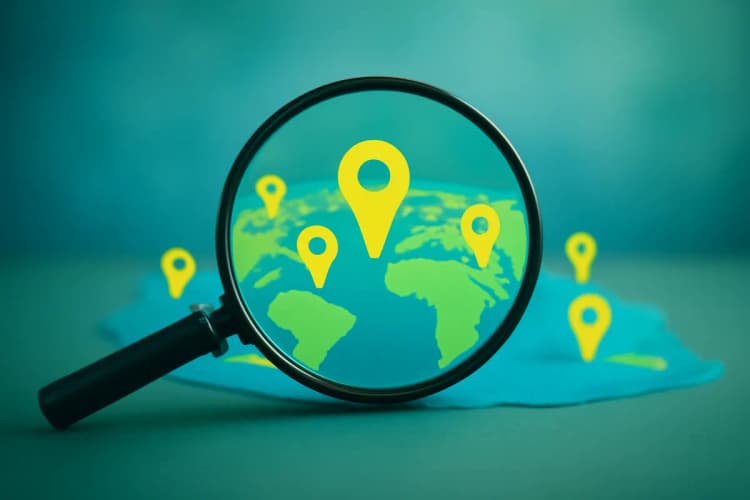 Pequena lupa sobre um mapa estilizado do mundo, com vários pinos de localização espalhados. A imagem é um detalhe, com fundo desfocado, em tons de azul e verde, transmitindo a ideia de alcance e visibilidade.